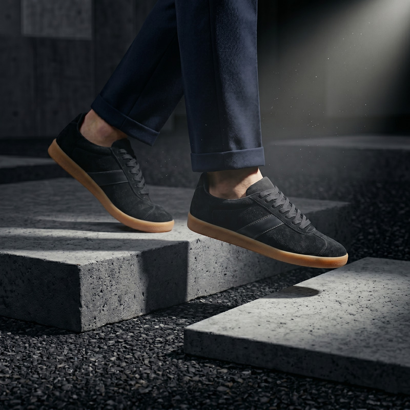 A person walking down concrete stairs wearing Asphalt black suede sneakers with gum soles and dark trousers.