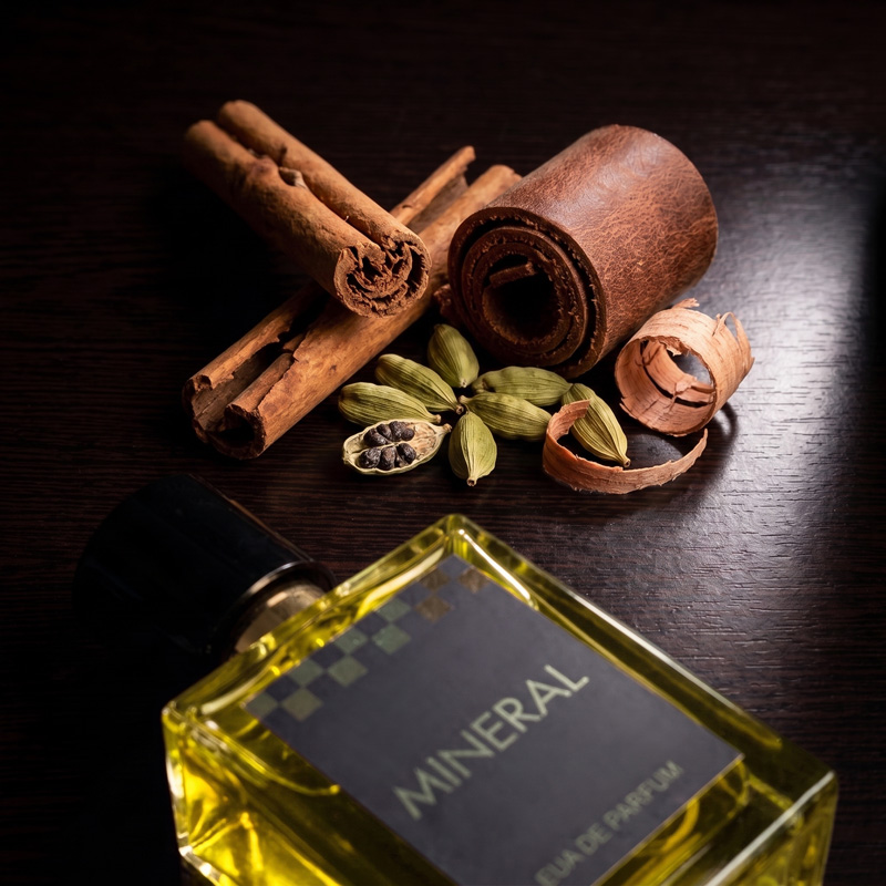 Mineral warm spicy perfume bottle next to raw fragrance ingredients including cinnamon sticks, green cardamom pods, and a rolled strip of brown leather